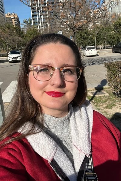 Cristina con una chaqueta roja y un peinado recogido, disfrutando de un día soleado mientras sonríe frente a la cámara en un parque urbano.Cristina con una chaqueta roja y un peinado recogido, disfrutando de un día soleado mientras sonríe frente a la cámara en un parque urbano.