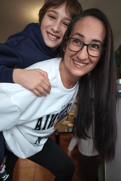 Cristina Asensio Aranda sonriendo mientras su hijo pequeño está sobre su espalda, ambos luciendo ropa cómoda y casual.