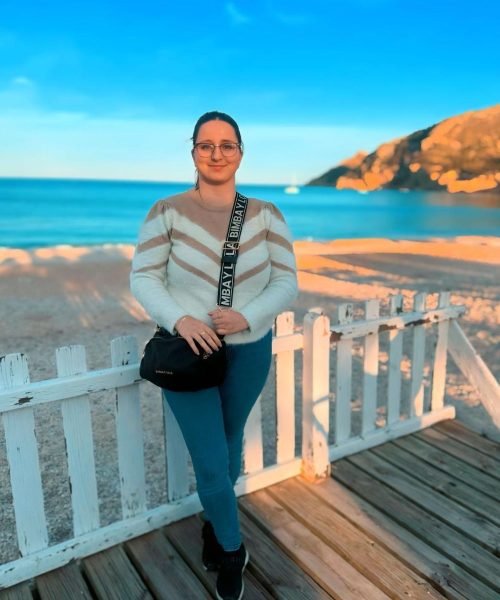 Cristina Pérez, creadora de contenido UGC, posando con suéter blanco, jeans azules y bolso negro en la playa, disfrutando de un paisaje al atardecer.