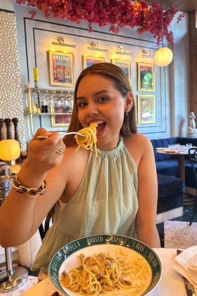 Mujer comiendo pasta en un restaurante con fondo decorado de flores y carteles.