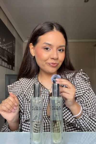 Mujer mostrando dos botellas de perfume mientras sostiene un aplicador con un gesto confiado.
