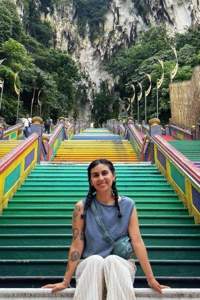 Fátima del Pilar sentada en las coloridas escaleras de Batu Caves en Malasia, rodeada de vegetación tropical.
