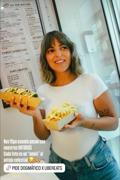 Natalia Márquez Sánchez sonriendo mientras sostiene dos bandejas con perritos calientes con salsas frente a un menú impreso en la pared.