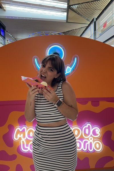 Natalia Márquez Sánchez posando divertida con un top y falda a rayas blancas y negras mientras muerde un perrito caliente frente a un fondo rosa y naranja.