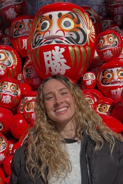 Creadora de contenido sonriendo frente a una colección de figuras Daruma tradicionales durante un viaje por Japón.