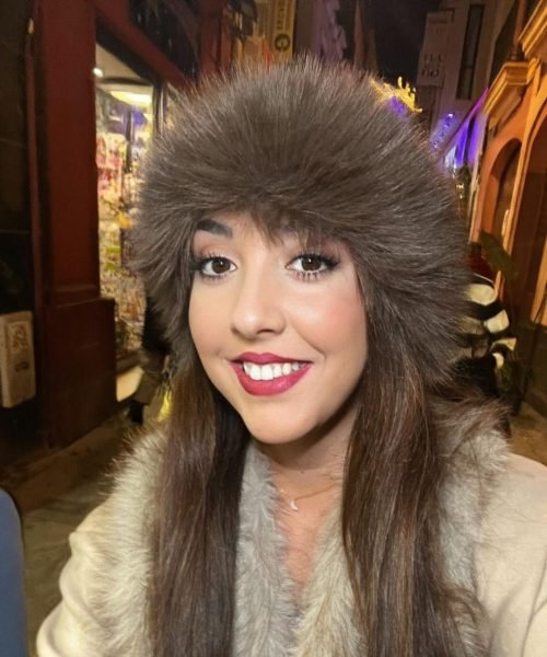 Lola Martín González con un gorro de piel sintética y abrigo blanco, sonriendo en una calle iluminada por luces nocturnas