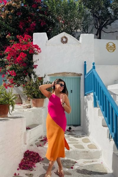 Lucía Pérez García luciendo un vestido bicolor rosa y naranja en una calle de arquitectura mediterránea con flores y escaleras blancas.