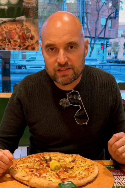 Hombre comiendo pizza en un restaurante, con una pizza frente a él y un cartel promocional al fondo.