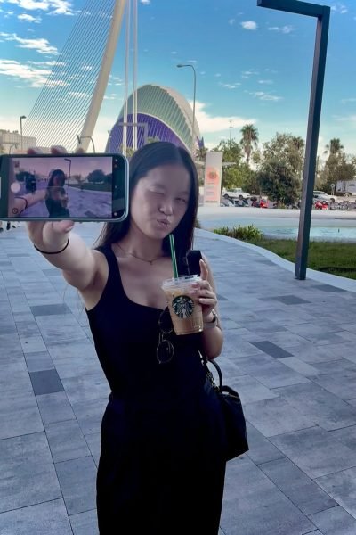 Agencia de marketing digital Brilla Influencers y Comunicación, María Bedins con vaso de Starbucks y teléfono móvil, posando frente a un fondo urbano con arquitectura moderna.