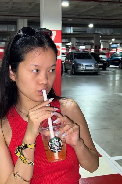Agencia de marketing digital Brilla Influencers y Comunicación, María Bedins con camiseta roja, sosteniendo vaso de Starbucks en estacionamiento.