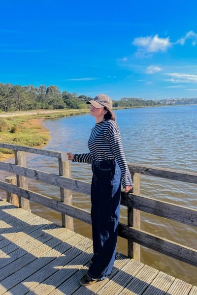 María Huidobro paseando por una pasarela de madera junto a una laguna, con outfit casual de rayas y gorra, disfrutando de un entorno natural y relajado.