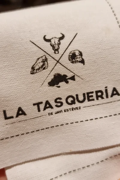 Logo de La Tasquería de Javi Estévez en un paño blanco, mostrando los íconos de la marca. Title: La Tasquería de Javi Estévez