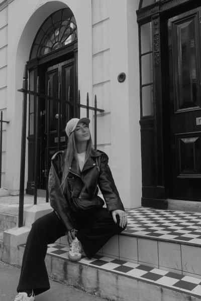 Mireya Sánchez sentada en la entrada de un edificio con chaqueta de cuero negra y gorra beige.