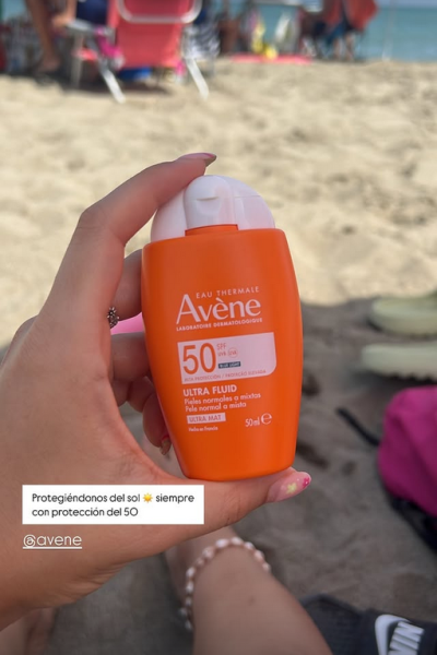 Protector solar Avène Ultra Fluid SPF 50 sostenido en la mano en un entorno de playa con sombrillas de fondo.