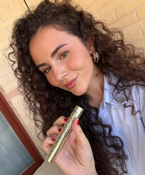 Agencia de marketing digital Brilla Influencers y Comunicación, mujer con rizos, usando barra de labios dorada, fondo de pared de ladrillos.