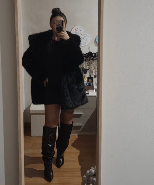 Veronica Villegas con abrigo de pelo negro, vestido corto y botas largas, posando frente al espejo.