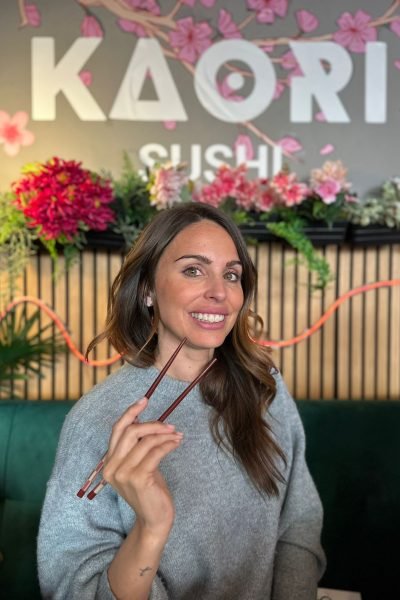 Mujer sonriendo mientras sostiene palillos frente al logo de Kaori Sushi.