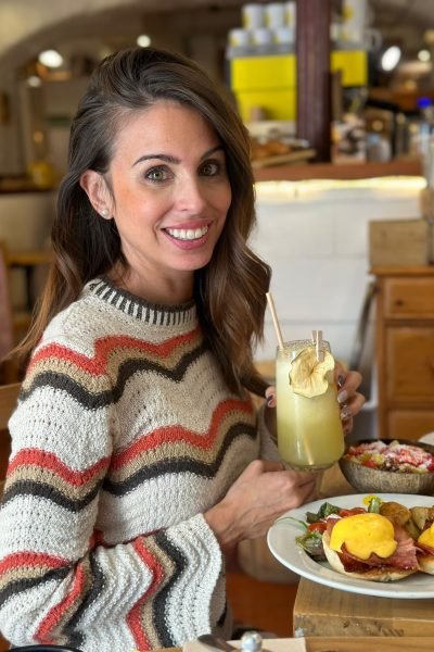 Mujer sonriendo mientras sostiene un vaso de jugo y un plato con comida, en un ambiente de café.