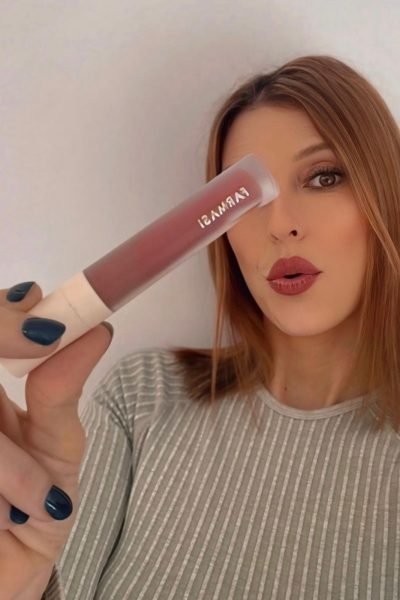 Mujer joven mostrando un labial líquido marrón sobre su ojo, con camiseta de rayas gris.
