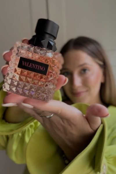 Mujer mostrando un frasco de perfume Valentino en primer plano, con una blusa verde brillante.