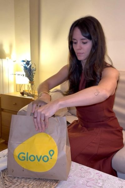 Esther Arribas Checa sentada en un sofá abriendo una bolsa de papel marrón con el logotipo amarillo y azul de Glovo sobre una mesa.