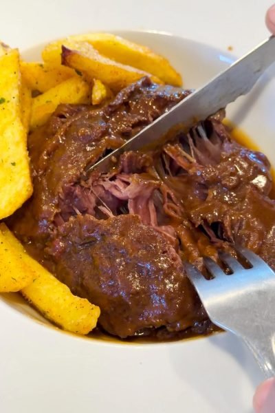 Croqueta recién frita abierta mostrando su relleno cremoso con patatas fritas al fondo, detalle foodie.
