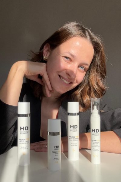 Alexandra Suciu mostrando los productos Melan TXA de HD Cosmetics, sonriendo mientras sostiene el sérum y las cremas para el cuidado de la piel.