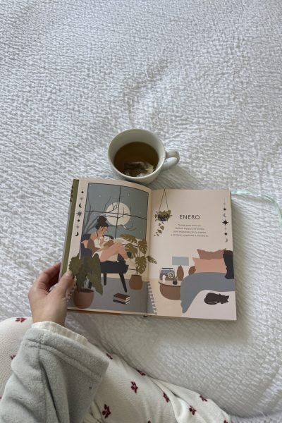 Mano sosteniendo un libro abierto con una ilustración, junto a una taza de té sobre manta blanca.