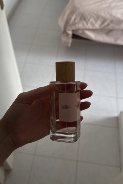 Mano sosteniendo perfume "Floral Breeze" con fondo neutro y cama de fondo.
