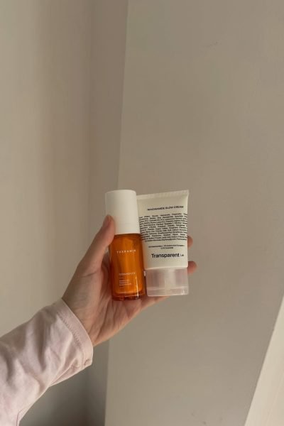 Productos de cuidado facial, serum y crema, en una mano frente a pared suave.