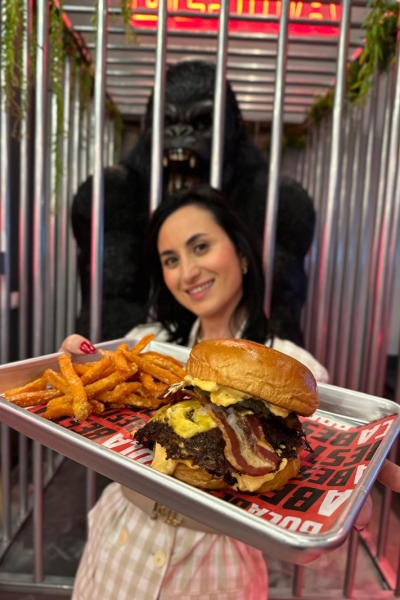 Laura Lobato sosteniendo una hamburguesa con bacon y patatas fritas en un restaurante de Madrid, sonriendo a cámara.