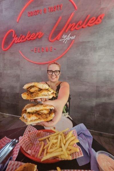 Marina García mostrando un sándwich y papas fritas en un restaurante, disfrutando de la comida y sonriendo a cámara.