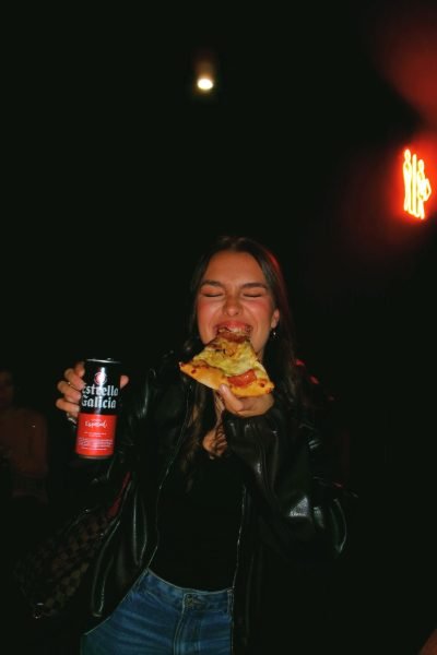 Marina Martínez sonriendo mientras sostiene una porción de pizza y una lata de cerveza Estrella Galicia en un ambiente nocturno.
