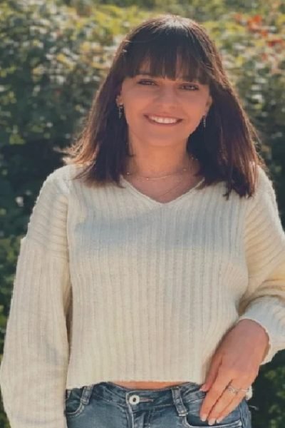 Mujer joven con jersey blanco y jeans, posando sonriente al aire libre frente a vegetación.